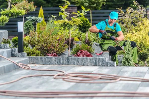 Parlak yeşillikler giymiş bir bahçıvan ve mavi bir kep dikkatlice budanmış çalılar ve bitkiler güpegündüz bakımlı bir bahçede. Bahçede çeşitli renkli bitkiler var..