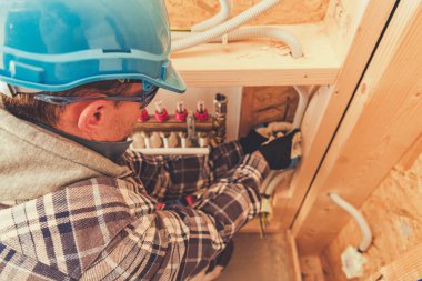 Mavi kasketli ve ekose gömlekli bir işçi inşaat halindeki yeni bir binaya tesisat parçaları yerleştiriyor..