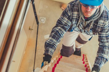 Bir inşaat işçisi güvenlik teçhizatı ve iş kıyafetleri giyerken tahta bir merdivene tırmanıyor ve gündüzleri tadilat alanındaki görevlerini tamamlamaya odaklanıyor..