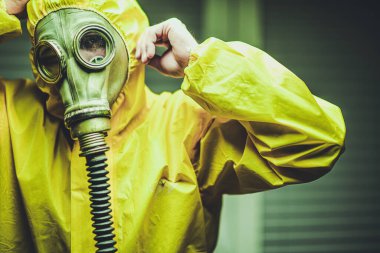 Canlı sarı koruyucu elbise giyen şahıs, acil durum tatbikatı sırasında güvenlik önlemlerini vurgulayarak, kentsel bir ortamda gaz maskesi takmaya hazırlanıyor..
