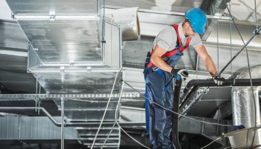 Bir HVAC çalışanı endüstriyel bir tesisin yukarısındaki havalandırma kanalını dikkatlice inceleyip onarıyor, güvenlik donanımı takıyor ve görevine odaklanıyor..