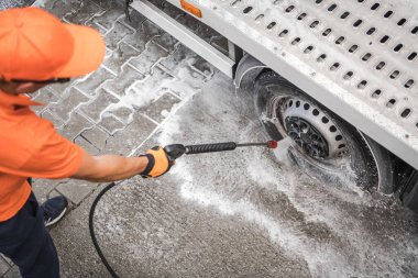 Turuncu şapkalı ve eldivenli bir işçi, temiz bir gökyüzünün altındaki kaldırımda sabun ve suyla çevrili bir kamyonun tekerleğini temizlemek için basınç makinası kullanır..