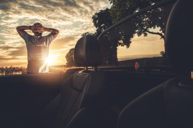 Bir adam arabasının dışında duruyor, sakin bir kırsalda canlı bir günbatımını seyrediyor. Gökyüzü sıcak renklerle dolu, rahatlatıcı bir atmosfer yaratıyor..