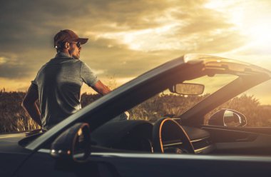 Bir adam üstü açık arabasının yanında oturur ve altın günbatımının tadını çıkarır. Huzurlu ufuk ve sıcak ışık, düşünmek için sakin bir atmosfer yaratır..