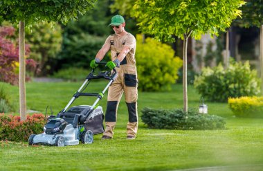 Profesyonel bir bahçıvan, güneşli bir öğleden sonra çalılar ve ağaçlarla dolu manikürlü bahçedeki canlı yeşil çimenleri budayarak bir çim biçme makinesi kullanır..