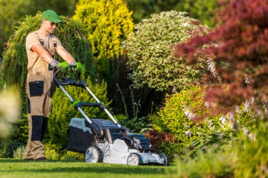 Bir bahçıvan yeşil çimenlikte çim biçme makinesini çalıştırıyor, çimenleri dikkatlice manikür ederken açık gökyüzünün altında renkli çalılar ve bitkilerle çevrili..