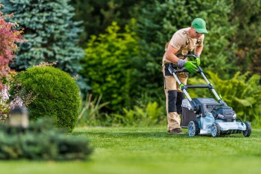 Etrafı çeşitli ağaçlar ve çalılarla çevriliyken bir adam güzel manzaralı bir bahçede çimleri biçiyor. Parlak güneş ışığı yeşilliği artırır..