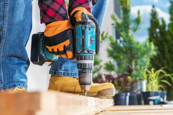 Bir insan açık havada ahşap işçiliğiyle meşgul, açık gökyüzünün altındaki tahtalar üzerinde elektrikli matkap kullanıyor. Güvenlik eldivenleri ve sağlam botlar koruma sağlar..