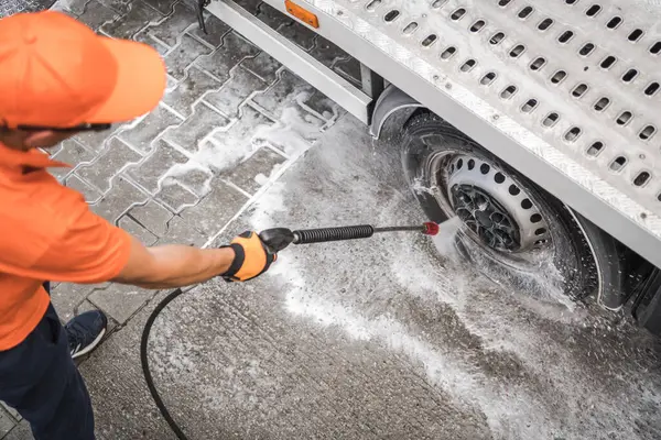 Turuncu şapkalı ve eldivenli bir işçi, temiz bir gökyüzünün altındaki kaldırımda sabun ve suyla çevrili bir kamyonun tekerleğini temizlemek için basınç makinası kullanır..