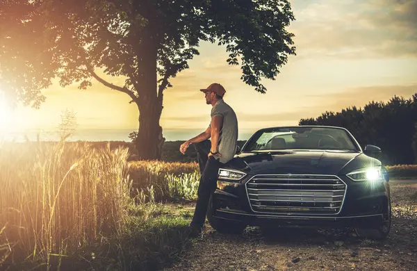Bir adam parlak siyah bir arabanın yanında duruyor, sakin bir kırsalda ağaçların ve tarlaların üzerinden canlı günbatımını seyrediyor..