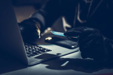 Koyu renk eldivenli bir kişi dizüstü bilgisayara odaklanır, yetersiz ışıklandırılmış bir alanda elinde bir kredi kartı tutuyordur, bu da sahte online davranışlar olduğunu gösterir..