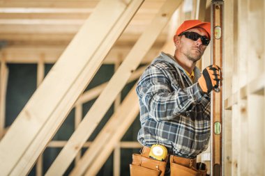 Yetenekli bir işçi, ahşap kirişi düzgün bir aletle ölçüyor ve bir çerçeve inşa ederken doğruluğu sağlıyor. Güvenlik teçhizatı ve odaklanmış ifade açık.