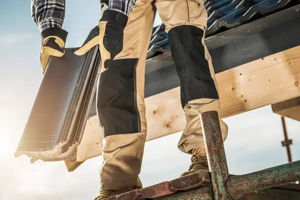 Becerikli bir çatı ustası iskelede duruyor, yerleşim yerlerinin çatısına fayanslar yerleştirmeye hazırlanıyor. Güneş ışıl ışıl parlıyor, verimli bir çalışma gününü işaret ediyor..