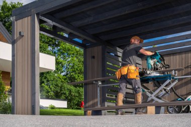 Bir marangoz, güneşli bir günde etrafı ağaçlarla çevrili bir açık arazinin altında elektrikli testereyle tahta kesmeye odaklanır..