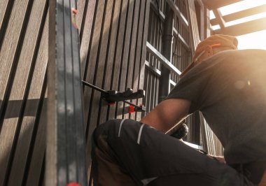 Kendini adamış bir zanaatkar, günbatımının sıcak ışıklarıyla güçlendirilmiş, çağdaş bir dış mekâna ahşap lamella çıtçıtası yerleştirmeye odaklanır. Görevde hassasiyet ve beceri açıkça görülür..