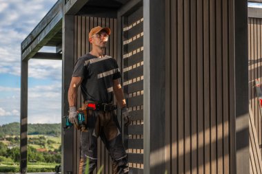 Yetenekli bir işçi, parlak mavi gökyüzünün altında yemyeşil bir manzarayla çevrili kısmen bitmiş ahşap bir yapının yanında duruyor. Aletler onun yanında..