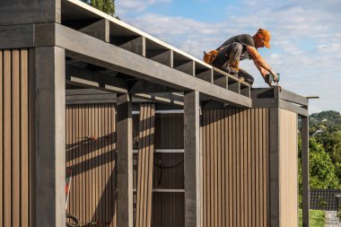 Becerikli bir işçi, verimli yeşillikler ve temiz bir gökyüzü ile çevrili iken ahşap panelleri dikkatlice yapıştırarak gösterişli bir baraka inşa ediyor..
