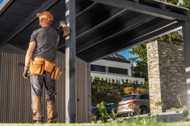 Bir inşaat işçisi garajın altında duruyor, eldiven ve alet kemeri takıp binanın bütünlüğünü kontrol ediyor. Güneş ışığı modern evi aydınlatır..