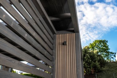 Güneş ışığı, verimli yeşillik ve parlak gökyüzü ile çevrili modern bir yapının parlak tahta levhalarına gölge düşürür..