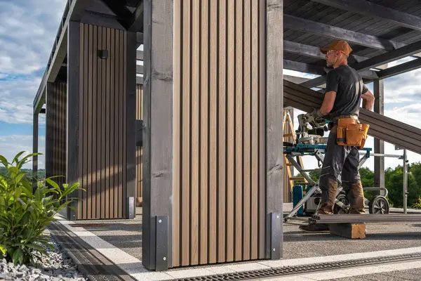 Bir marangoz, aletler ve malzemelerle çevrili, açık hava alanındaki ahşap bir yapı üzerinde özenle çalışır..