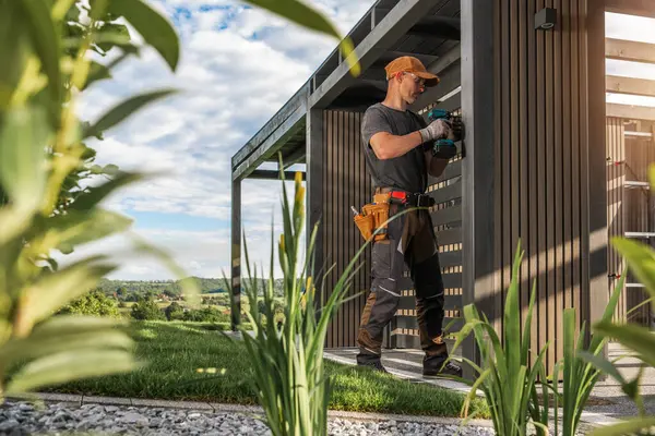 Bir inşaat işçisi, yeşillik ve açık gökyüzü ile çevrili şık bir açık hava yapısına elementleri dikkatlice yerleştirir. Canlı bahçe ferahlatıcı bir dokunuş ekliyor..