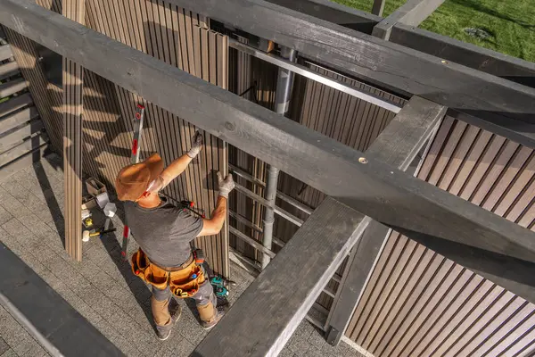Bir inşaat işçisi, açık mavi gökyüzünün altındaki bir konut projesinde tahta kirişleri güvence altına almak için merdiven kullanıyor. Çalışma alanı iyi organize edilmiş ve verimli..