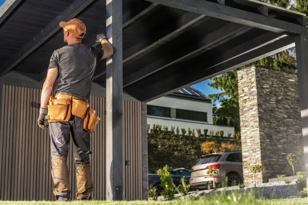 Bir inşaat işçisi garajın altında duruyor, eldiven ve alet kemeri takıp binanın bütünlüğünü kontrol ediyor. Güneş ışığı modern evi aydınlatır..