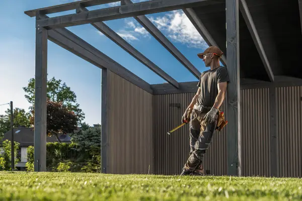Bir inşaat işçisi, elinde aletlerle, güneşli bir günde çevresi yeşilliklerle çevrili bir bina yapısının gelişimini güvenli bir şekilde izler..