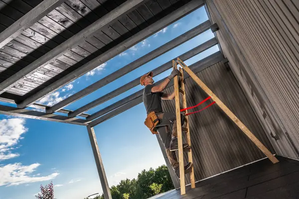 Bir işçi, çağdaş bir çardağın tavan panellerini dikkatlice yerleştirmek için merdiven kullanıyor. Parlak mavi gökyüzü parlak öğleden sonra ışığını yansıtıyor, sahneyi güçlendiriyor..