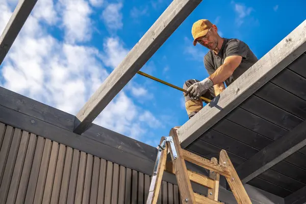 Bir inşaat işçisi parlak mavi bir gökyüzünün altındaki merdivene tünemiş ahşap kirişleri ölçmeye odaklanır. Bir yenileme projesi üzerinde çalışıyor..