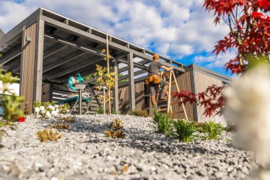 Bir inşaat işçisi ahşap bir yapıyı bir araya getirmek için merdiveni tırmanır güzel, mavi gökyüzünün altında, kabarık bulutlu bir bahçe..