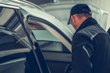 Siyah ceketli bir tamirci garajın içindeki gümüş bir araca yaklaşır ve arabaya girmeye odaklanır. Güneş ışığı filtreler, boşluğu aydınlatır.