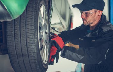 Tamirci, iyi aydınlatılmış bir oto tamirhanesinde bir araba lastiğini inceler ve tamir eder..