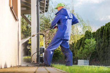 Mavi üniformalı bir işçi evin dışını temizlemek için elektrikli çamaşır makinesi kullanır. Güneşli bir gün ve arka planda yeşil ağaçlar görünüyor..