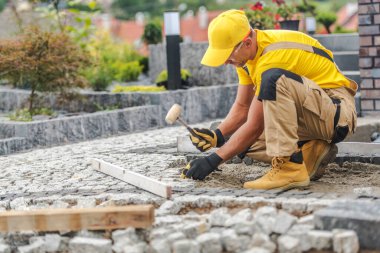Bir inşaat işçisi bahçeye kaldırım taşı koymaya odaklanır. Yeşil bitkiler ve renkli çiçeklerle çevrili her taşı özenle ayarlamak için bir çekiç kullanıyor..