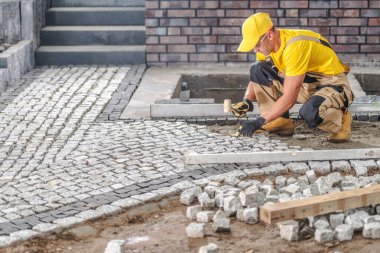 Sarı şapkalı ve gömlekli bir işçi gün ışığında inşaat alanındaki kaldırım taşlarını dikkatlice yere yerleştirir..