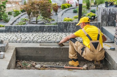 Sarı tişörtlü ve şapkalı bir işçi yeşil yollarla çevrili bir bahçe yolunda taşların düzgün hizalanmasını sağlamak için bir seviyelendirme aracı kullanıyor..