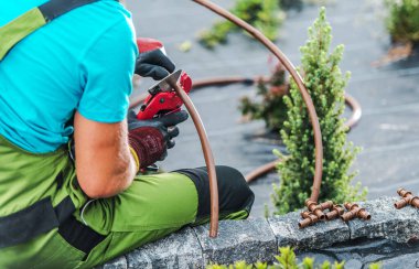 Bir bahçıvan peyzaj alanındaki sulama borularını kesmeye odaklanır. Çevreleyen bitkiler açık havada gün ışığında çalışkan çalışmaları vurguluyor..