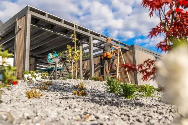 Bir inşaat işçisi ahşap bir yapıyı bir araya getirmek için merdiveni tırmanır güzel, mavi gökyüzünün altında, kabarık bulutlu bir bahçe..