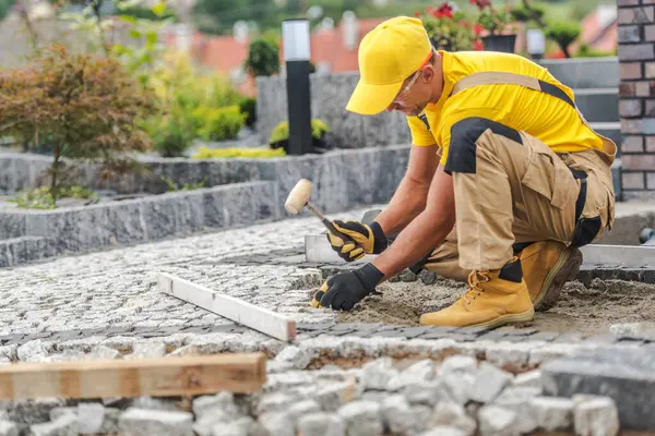 Bir inşaat işçisi bahçeye kaldırım taşı koymaya odaklanır. Yeşil bitkiler ve renkli çiçeklerle çevrili her taşı özenle ayarlamak için bir çekiç kullanıyor..