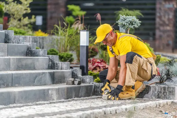 Sarı tişörtlü ve şapkalı bir işçi, taşları dikkatlice bir dizi açık hava basamağına yerleştiriyor. Yakınlardaki bitkiler ve dekorasyonlar çevreyi güzelleştirir, canlı bir sahne yaratır..