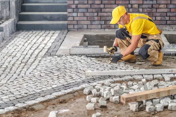Sarı şapkalı ve gömlekli bir işçi gün ışığında inşaat alanındaki kaldırım taşlarını dikkatlice yere yerleştirir..