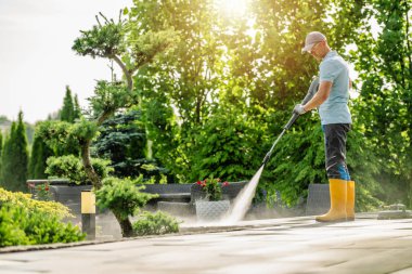 Mavi gömlekli ve sarı çizmeli bir adam terası temizliyor. Yeşillik onu çevrelemiş, canlı bir açık hava atmosferi yaratmış..
