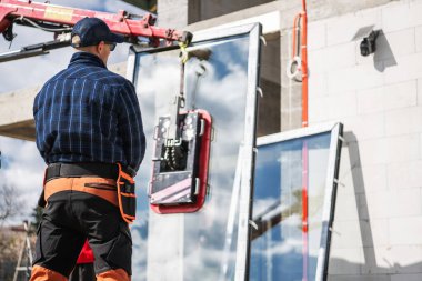 Koruyucu ekipman işçisi, bir inşaat alanındaki büyük cam panelleri kaldırıp güvenlik ve hassasiyeti sağlayan bir vinci denetliyor..