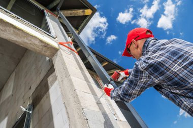 Bir inşaat işçisi güneşli bir günde binanın dış duvarına malzeme uyguluyor. İşçi işine odaklanmış, koruyucu giysiler giyiyor..