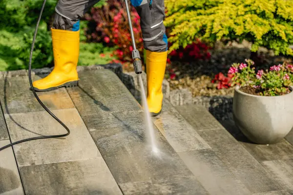 Sarı lastik çizme giyen biri, taş bir verandada basınçlı çamaşır makinesi kullanır. Renkli çiçekler ve yeşillik parlak, davetkar bir bahçe atmosferi yaratır..