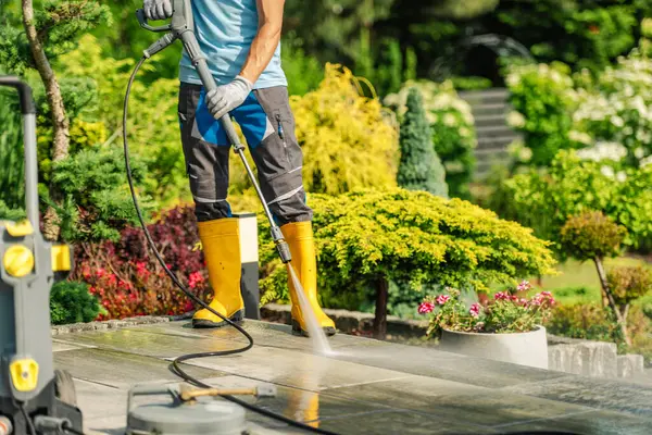 Sarı çizmeli bir işçi, canlı bitkiler ve çiçeklerle çevrili bir teras alanını temizlemek için basınç makinası kullanıyor. Bahçede güneşli havanın tadını çıkarıyor..