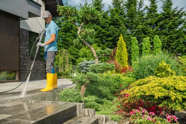 Sarı çizmeli bir adam, güneşli bir günde çevrenin canlı yeşillik ve çalılarla çevrili olduğu taştan bir verandayı temizlemek için çamaşır makinesi kullanıyor..