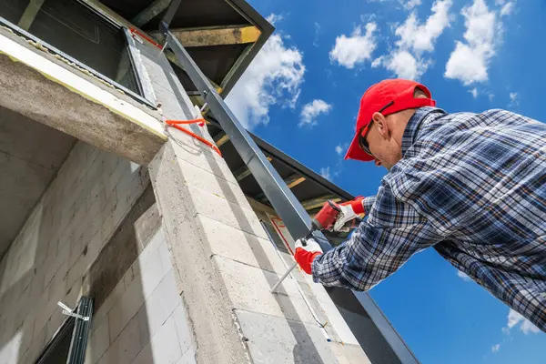 Bir inşaat işçisi güneşli bir günde binanın dış duvarına malzeme uyguluyor. İşçi işine odaklanmış, koruyucu giysiler giyiyor..