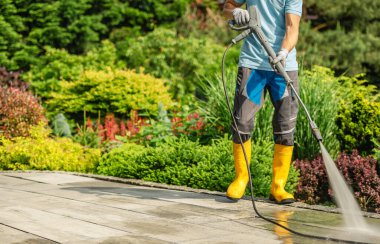 Sarı çizmeli bir bahçıvan güneşli bir yaz gününde, canlı bitkiler ve çiçeklerle çevrili bir terası basınç makinesiyle temizler..